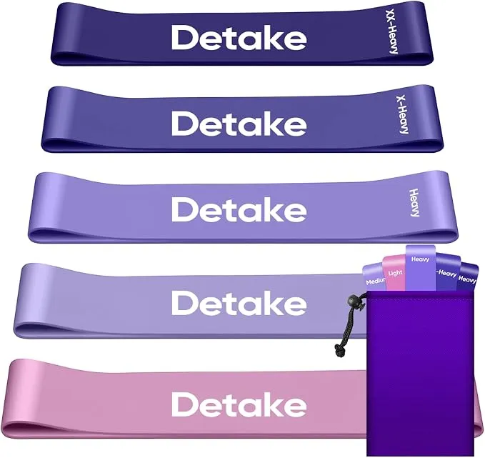 Cinq mini-bandes de résistance Detake de couleurs dégradées (violet foncé à rose clair), empilées, avec leurs niveaux de résistance inscrits (XX-Heavy, X-Heavy, Heavy, Medium, Light). Un petit sac de rangement violet est visible sur le côté droit.