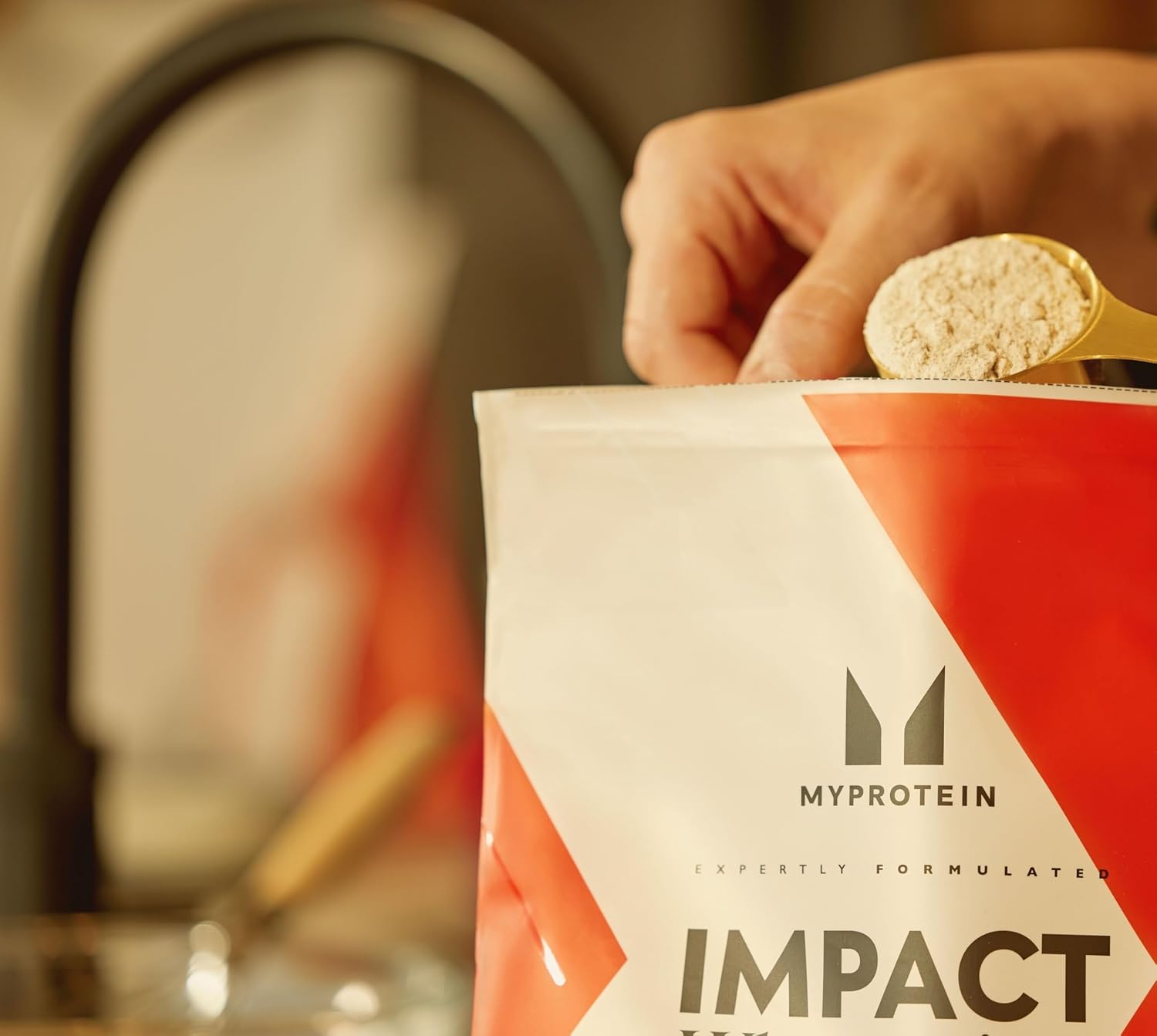Une main tenant une cuillère dorée remplie de poudre de gainer, sur le point d'être versée dans le sac de Myprotein Impact Whey Gainer, avec le haut du sac visible.